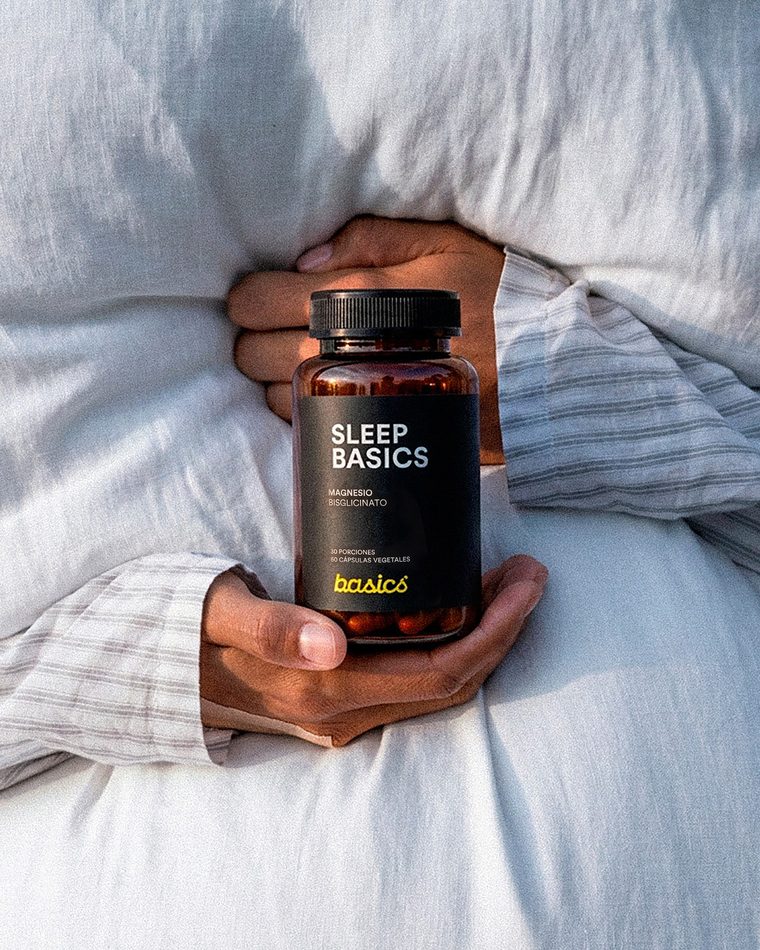 Pieza de Basics después: manos sosteniendo un frasco de Sleep Basics entre sábanas blancas